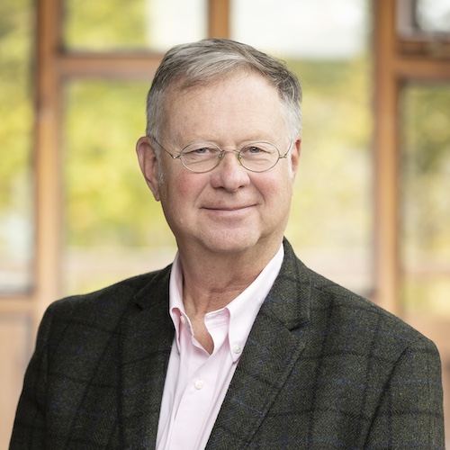 A headshot of Bruce Lourie wearing a grey blazer. A blurred background with photos of trees are behind him.
