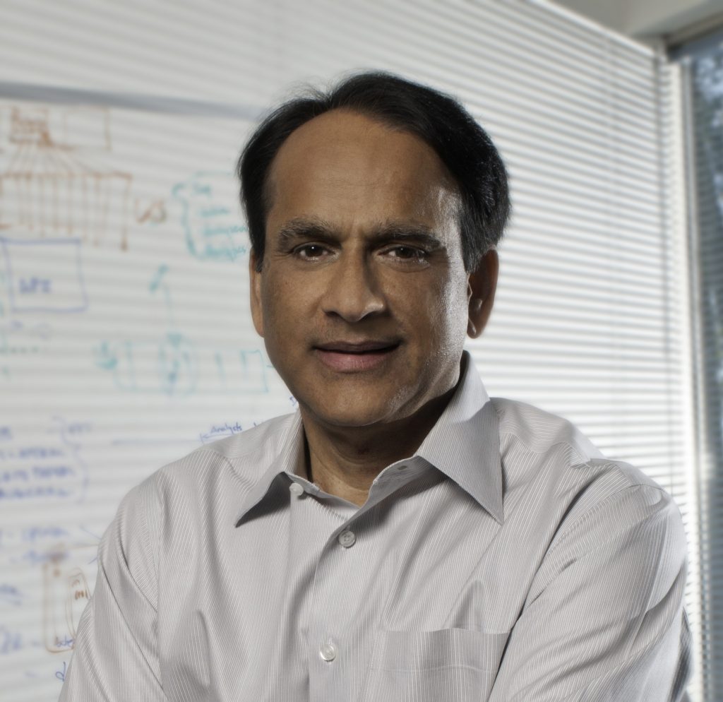 A headshot of Hassan Ahmed wearing a white collared shirt. A whiteboard behind him displays words and drawings in coloured marker.